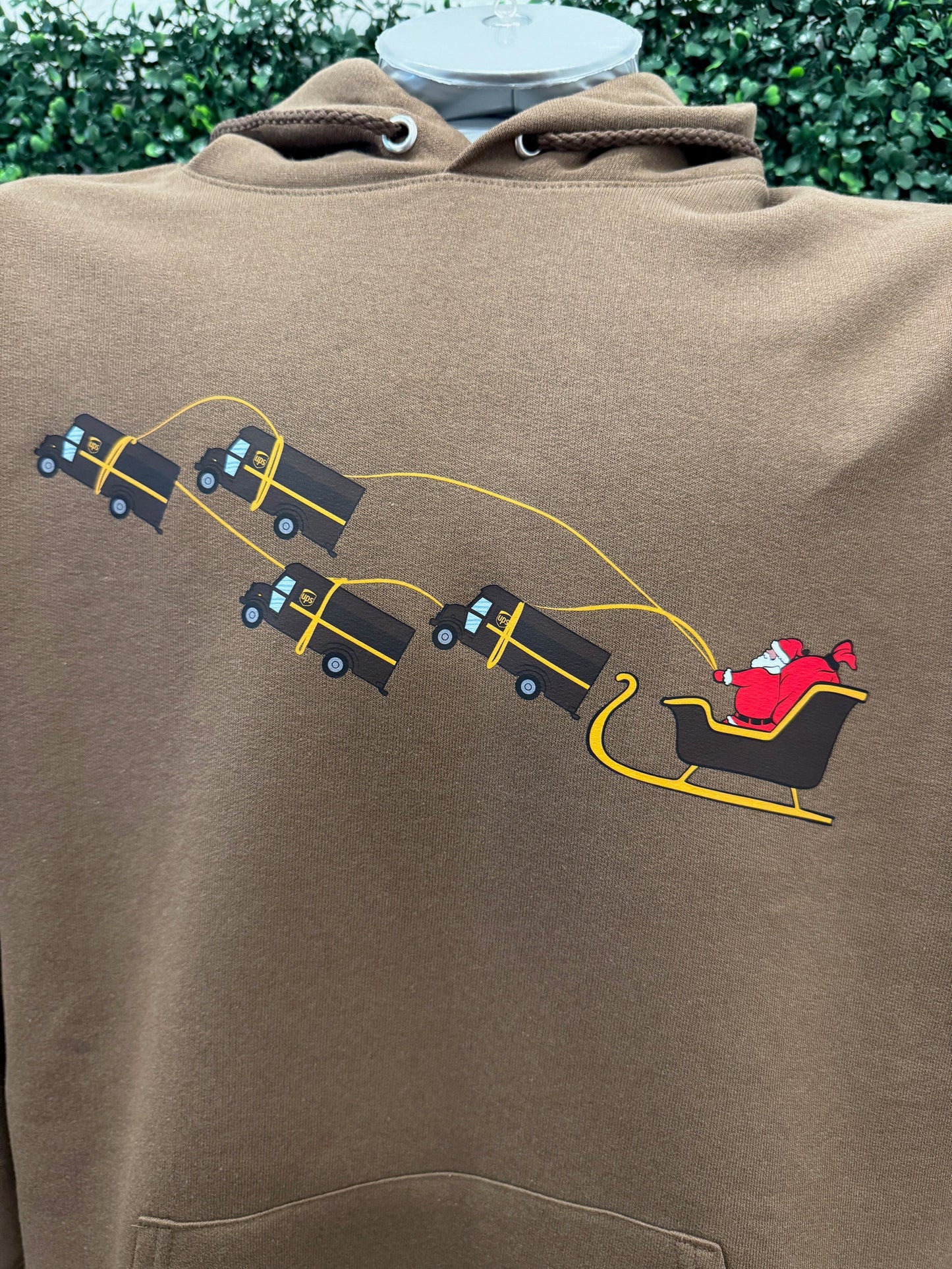 UPS Santa Peak Season Christmas Hoodie featuring Santa in a sleigh pulled by UPS trucks on brown fabric.