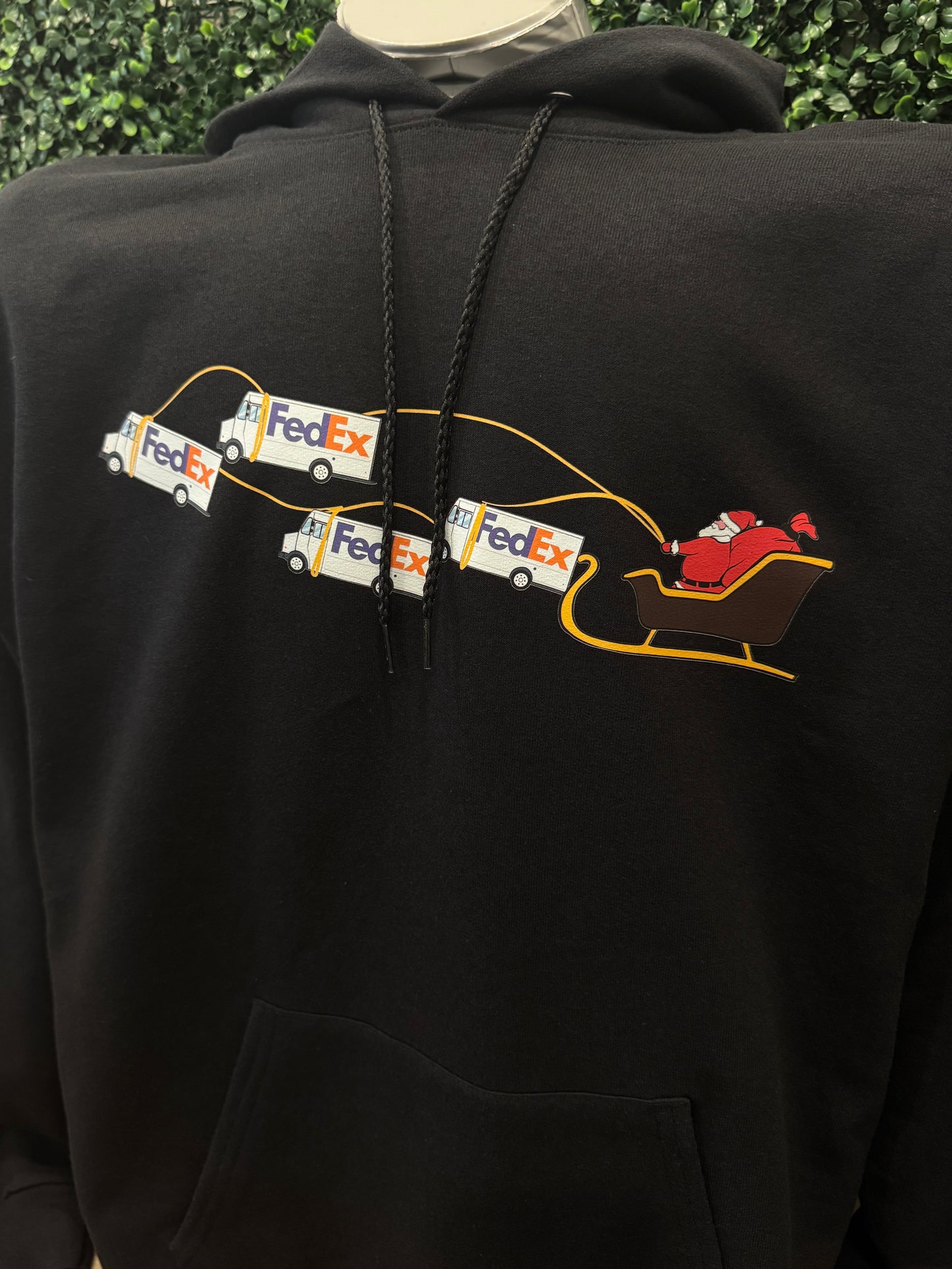 Black hoodie with FedEx logo and Santa Claus sleigh design on the back against a green leafy background.